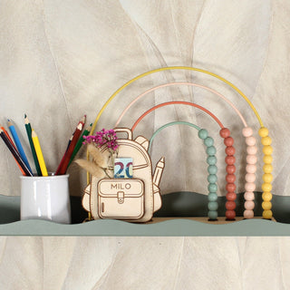 Personalisiertes Geschenk zur Einschulung aus Holz in Form einer Schultasche mit Stiften und Regenbogen auf einem Regal dekoriert