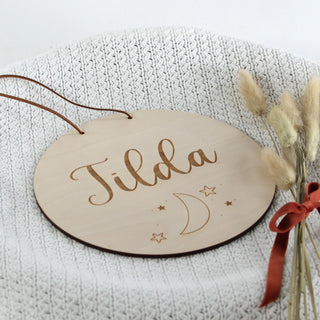 Rundes personalisiertes Namensschild aus Holz mit dem Namen, graviertem Mond- und Sternenmotiv, ansicht von oben – liebevolle Wanddeko fürs Kinderzimmer.