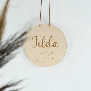 Rundes personalisiertes Namensschild aus Holz mit dem Namen, graviertem Mond- und Sternenmotiv, aufgehängt an einem Lederband – liebevolle Wanddeko fürs Kinderzimmer.