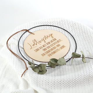 Rundes Holzschild mit schwarzem Metallrahmen und Spruch „Lieblingstage sind die mit dem Lachen…“ – handgefertigte Wanddeko aus Holz, liebevoll gestaltet für besondere Momente. uf weißer Strickdecke liegend.