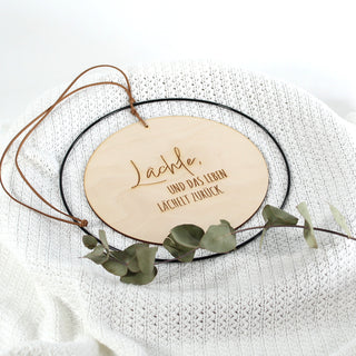 Rundes Holzschild mit dem Zitat „Lächle, und das Leben lächelt zurück.“ Eingefasst in einen schwarzen Metallring und aufgehängt an einem Kunstlederband – tolles Geschenk für viele Anlässe. Liegend auf weißer Strickdecke.