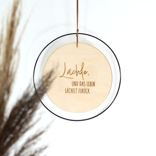 Rundes Holzschild mit dem Zitat „Lächle, und das Leben lächelt zurück.“ Eingefasst in einen schwarzen Metallring und aufgehängt an einem Kunstlederband – tolles Geschenk für viele Anlässe.
