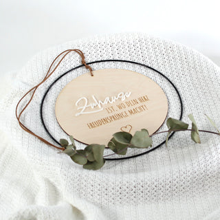 Rundes Holzschild mit dem Spruch „Zuhause ist, wo dein Herz Freudensprünge macht!“, schwarzer Metallrahmen, erhabenes Wort „Zuhause“ aus weißem Acryl, restlicher Text graviert. Auf weißer Strickdecke liegend.