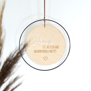 Rundes Holzschild mit dem Spruch „Zuhause ist, wo dein Herz Freudensprünge macht!“, schwarzer Metallrahmen, erhabenes Wort „Zuhause“ aus weißem Acryl, restlicher Text in schwarzer Vinylfolie, aufgehängt an einem Kunstlederband – moderne Wanddeko im natürlichen Stil.