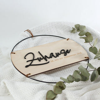 Rundes Holzschild mit dem Spruch „Zuhause ist, wo wir zusammen sind!“, schwarzer Metallrahmen, erhabenes Wort „Zuhause“ aus schwarzem MDF gelasert, restlicher Text in das Holz graviert, aufgehängt an einem Kunstlederband – moderne Wanddeko im natürlichen Stil. Ansicht von oben, dekoriert mit Eukalyptus auf einer weißen Strickdecke.