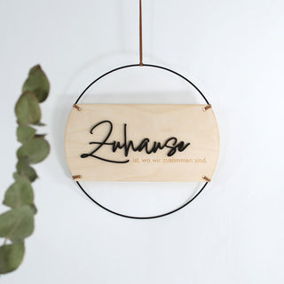 Rundes Holzschild mit dem Spruch „Zuhause ist, wo wir zusammen sind!“, schwarzer Metallrahmen, erhabenes Wort „Zuhause“ aus schwarzem MDF gelasert, restlicher Text in das Holz graviert, aufgehängt an einem Kunstlederband – moderne Wanddeko im natürlichen Stil.