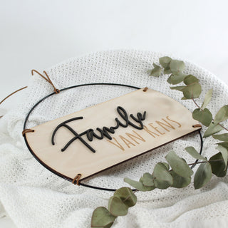 Personalisierbares Holzschild mit 3D-Schriftzug „Familie“ aus schwarzem MDF und lasergraviertem Nachnamen, eingefasst in einen schwarzen Metallring und mit Kunstlederband. Ideal als Wanddeko oder Türschild, handgefertigt und individuell gravierbar. Ansicht von oben auf Strickdecke mit Eukalyptus dekoriert. 