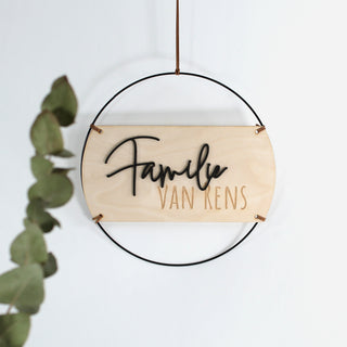 Personalisierbares Holzschild mit 3D-Schriftzug „Familie“ aus schwarzem MDF und lasergraviertem Nachnamen, eingefasst in einen schwarzen Metallring und mit Kunstlederband. Ideal als Wanddeko oder Türschild, handgefertigt und individuell gravierbar.