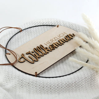 Handgefertigtes Holzschild mit dem Schriftzug „Herzlich Willkommen“, 3D-Schrift, filigrane Gravur auf hellem Holz, eingefasst in schwarzen Metallring, aufgehängt mit Kunstlederband – stilvolle Wand- und Türdeko.
