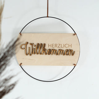 Handgefertigtes Holzschild mit dem Schriftzug „Herzlich Willkommen“, 3D-Schrift, filigrane Gravur auf hellem Holz, eingefasst in schwarzen Metallring, aufgehängt mit Kunstlederband – stilvolle Wand- und Türdeko.