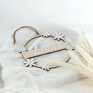 Personalisierter Namenskranz aus Holz mit weißen Gänseblümchen und 3D-Acryl-Schriftzug – handgemachte Kinderzimmer Deko mit Wunschname. Ansicht von oben auf weißer Strickdecke mit Pampasgras.