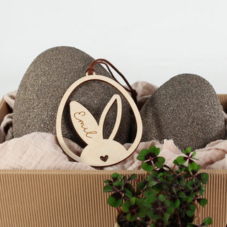 Personalisierter Osteranhänger aus Holz mit Hasenohren im Ei, gelasert mit Namen. Moderne Osterdekoration zum Aufhängen, dekoriert in einem Osterkörbchen.