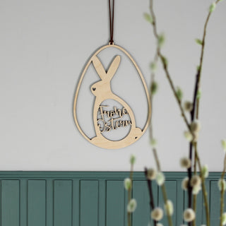 Holzschild in Osterei-Form mit Hasen-Silhouette und ausgelasertem Ei mit Schriftzug „Frohe Ostern“. Dekoriert über grüner Holzpaneele Wand.