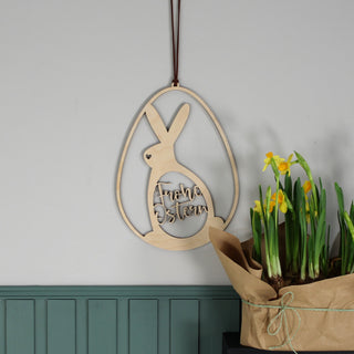 Holzschild in Osterei-Form mit Hasen-Silhouette und ausgelasertem Ei mit Schriftzug „Frohe Ostern“. Osterlich dekoriert über grüner Holzpaneele Wand.